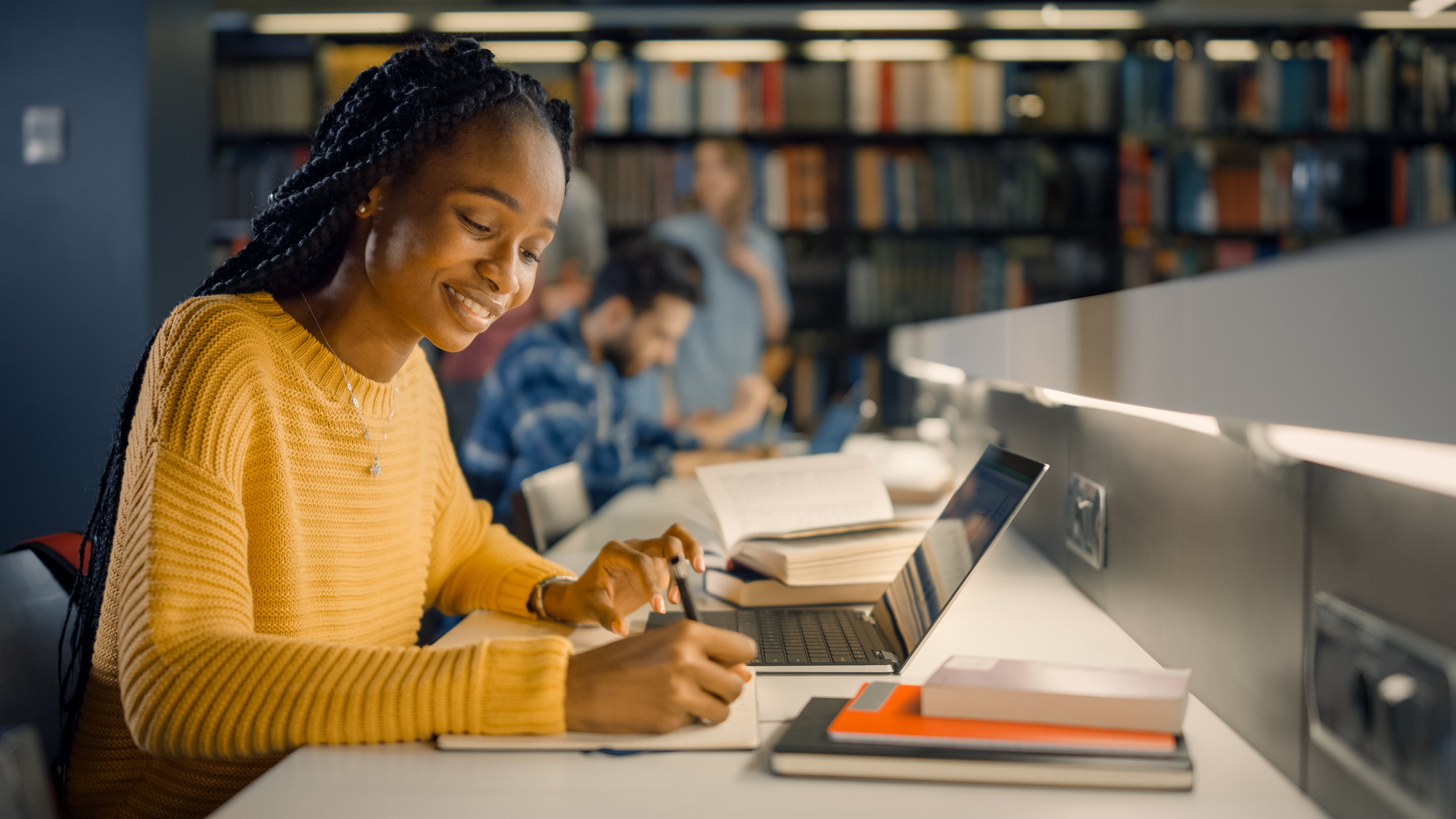 uma mulher universitária estudando usando um notebook em uma biblioteca. Ao fundo observamos algumas pessoas desfocadas.